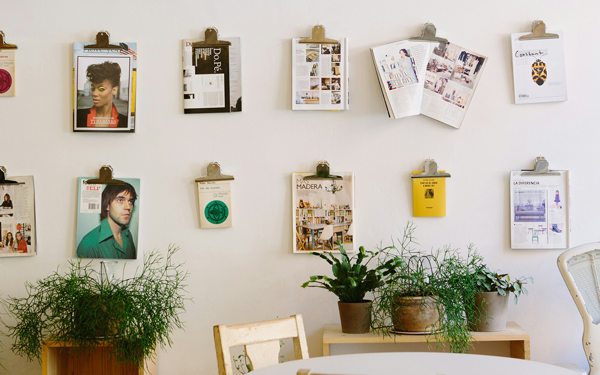 a variety of photos and art works pinned to a white wall, in two slightly askew rows, over a table with greenery on it.