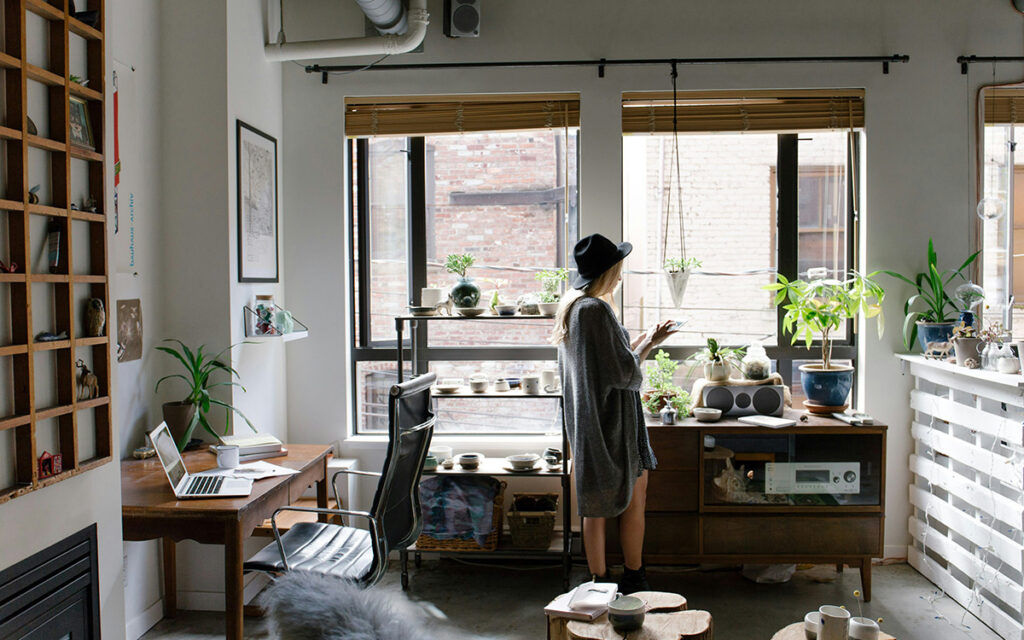 A female presenting person wearing a grey shirt dress and black felt hat stands in the centre of a home studio space holding an ipad. The room is organized and many art works are displayed on the various furniture pieces and walls.