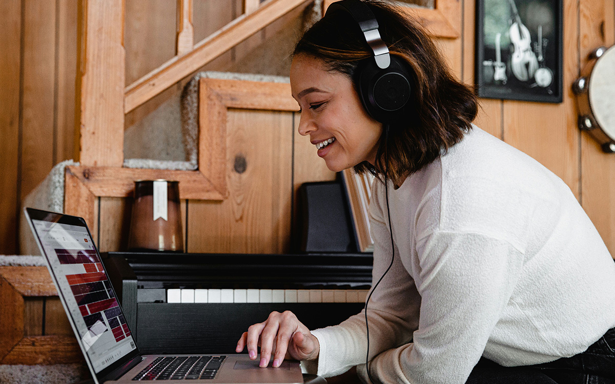 A female presenting person sits in a living room working on a laptop. They are wearing a white shirt and noise cancelling headphones.