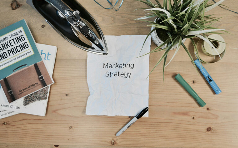 A photo taken from over top of a desk. There's a wrinkled paper on the desk with the works Marketing Strategy on it and a clothes iron resting on the corner of it. Various office supplies are scattered nearby.